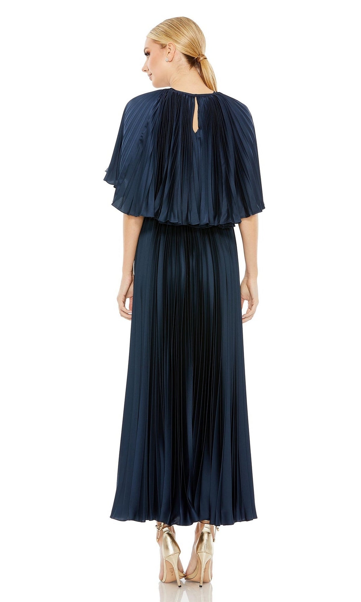 A woman faces away, wearing the Mac Duggal 49638 Caped Tea-Length Party Dress in navy blue with pleats and a cape-like overlay, paired with metallic high heels.